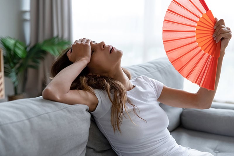 Why Is My AC Blowing Hot Air? Photo of an overheated woman fanning herself to cool off on her couch.